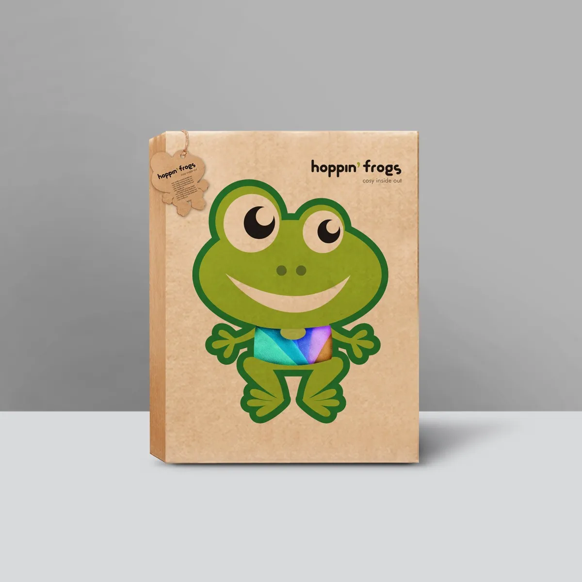hoppin' frogs kraft gift box — smiling cartoon frog mascot printed full height on natural brown cardboard, holding rainbow-coloured underwear visible through die-cut window; frog-shaped hang tag at left