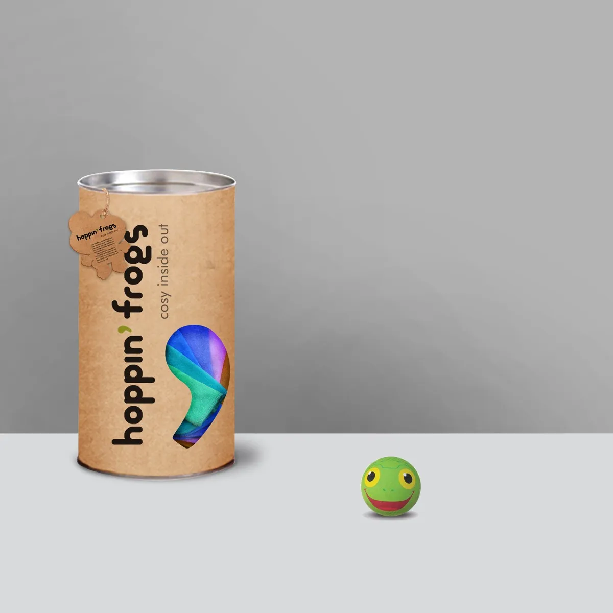 hoppin' frogs kraft cylinder tube — vertical wordmark with 'cosy inside out' tagline, foot-shaped die-cut window showing colourful socks; small green frog-face ball toy beside it