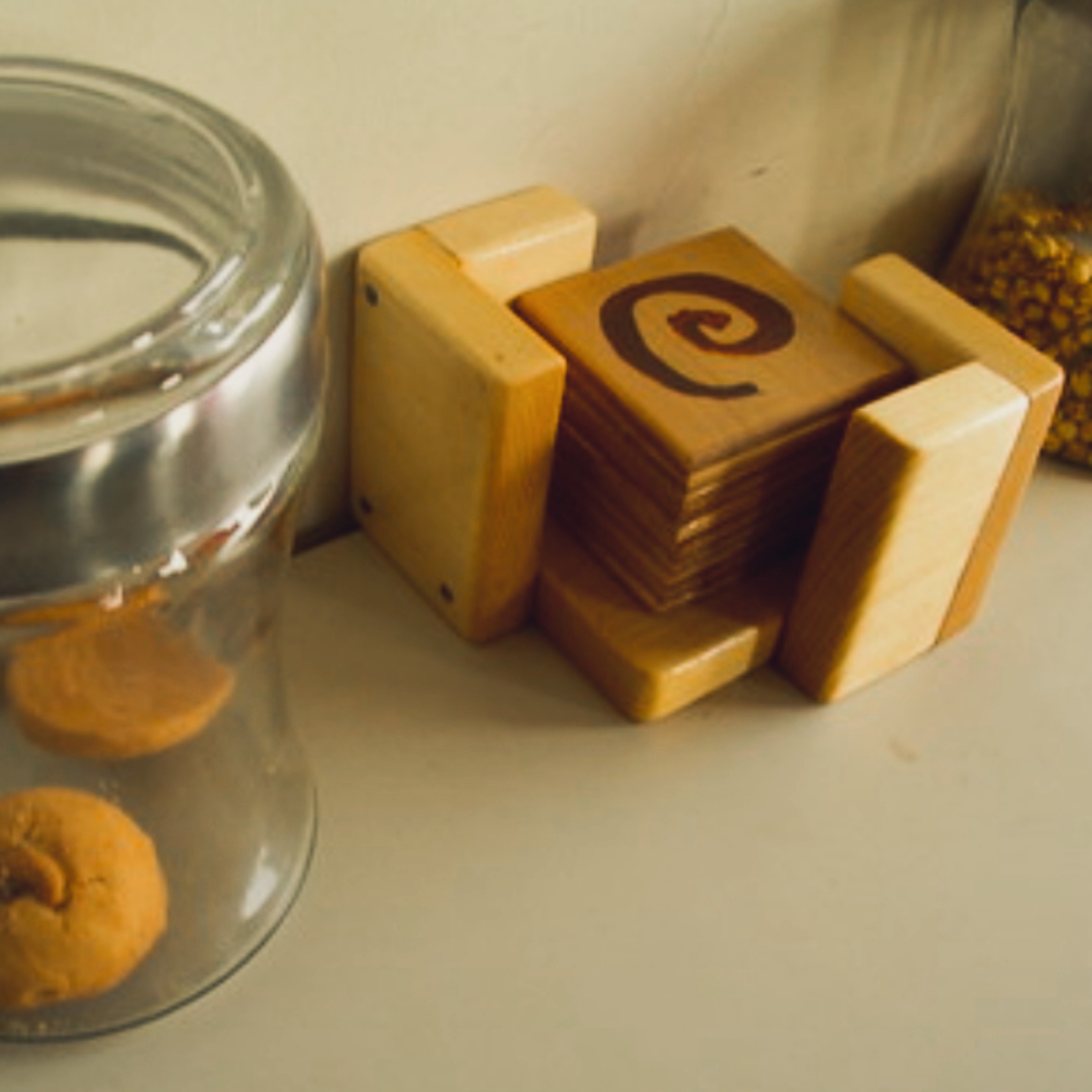 Set of hand-cut wooden square coasters with a spiral burn design on the top coaster, propped against a glass cookie jar