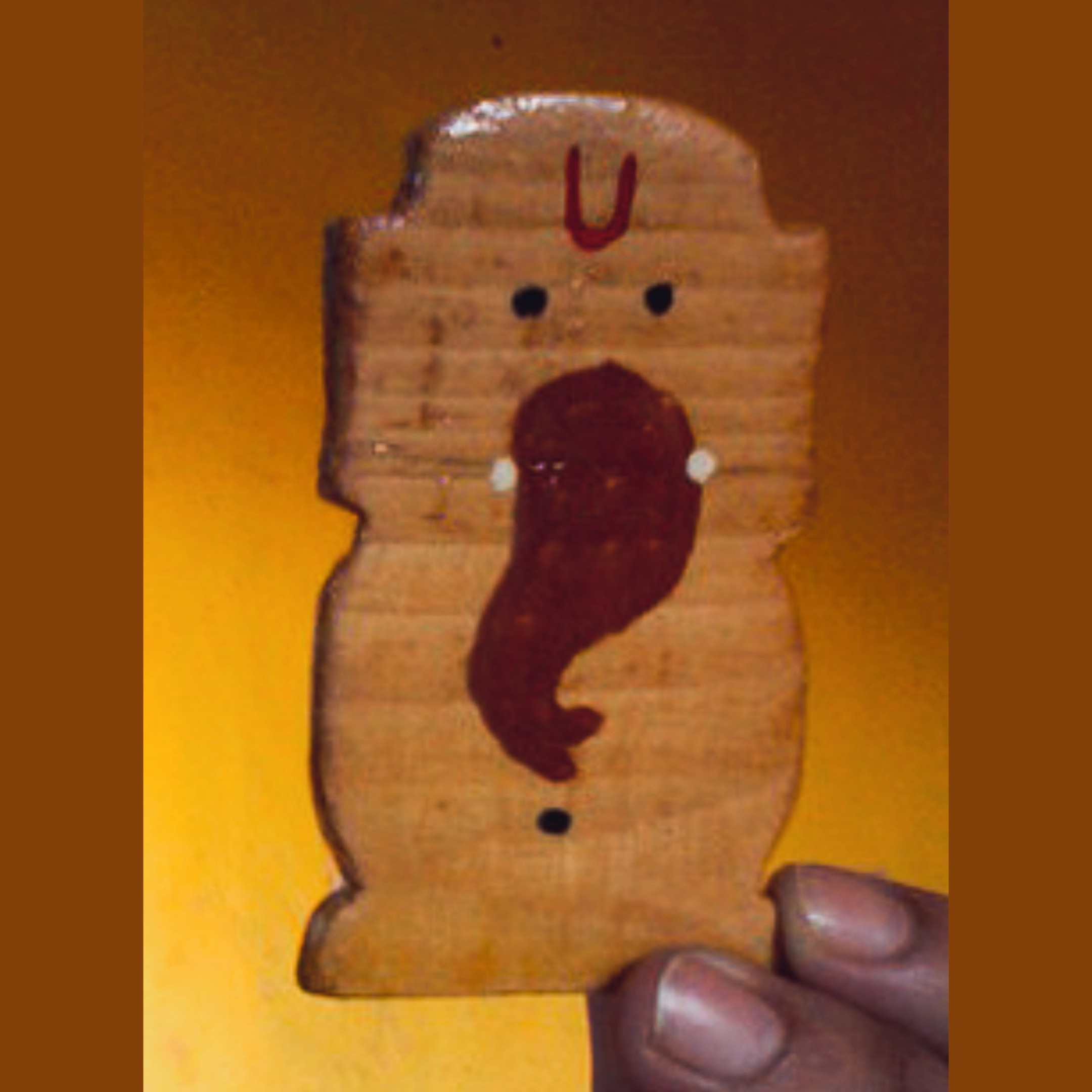 Tiny hand-carved wooden Ganesha figurine held in fingers — simplified abstract form with tilak U-mark in red