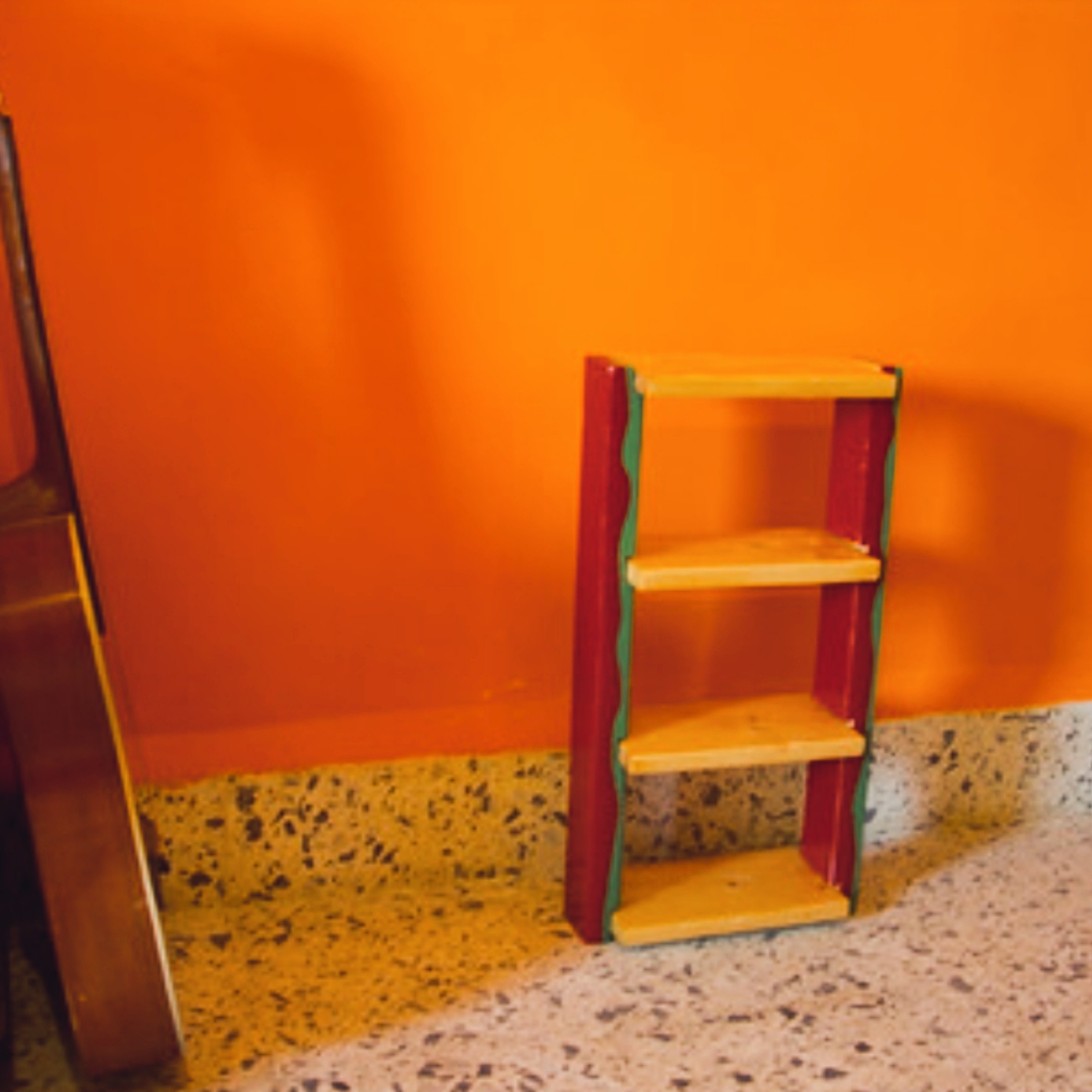 Small three-shelf unit with red sides and wavy painted edges in yellow and green, standing against an orange wall