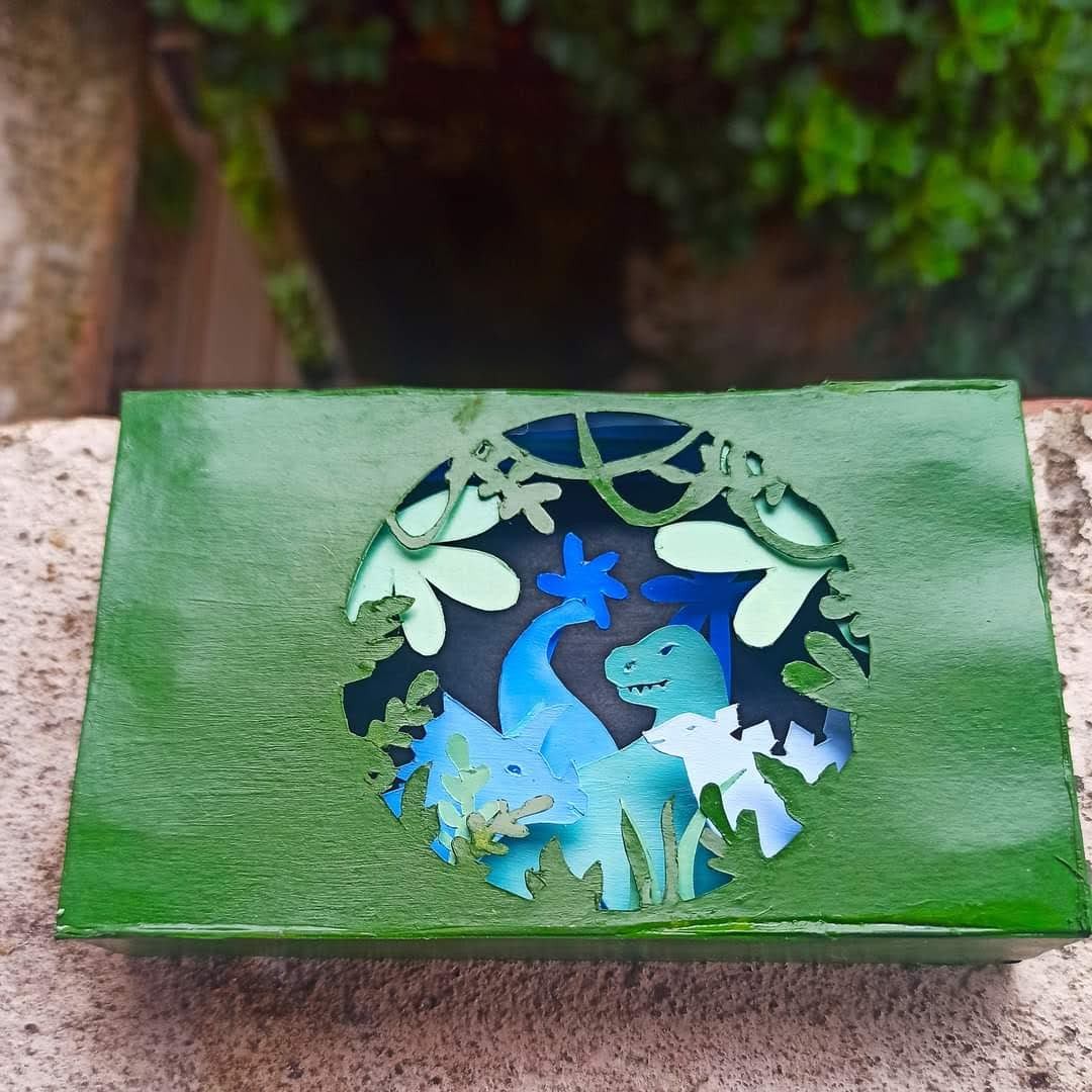 Layered cardboard dinosaur diorama inside a green Amazon box — T-rex and brachiosaurus in blue and teal