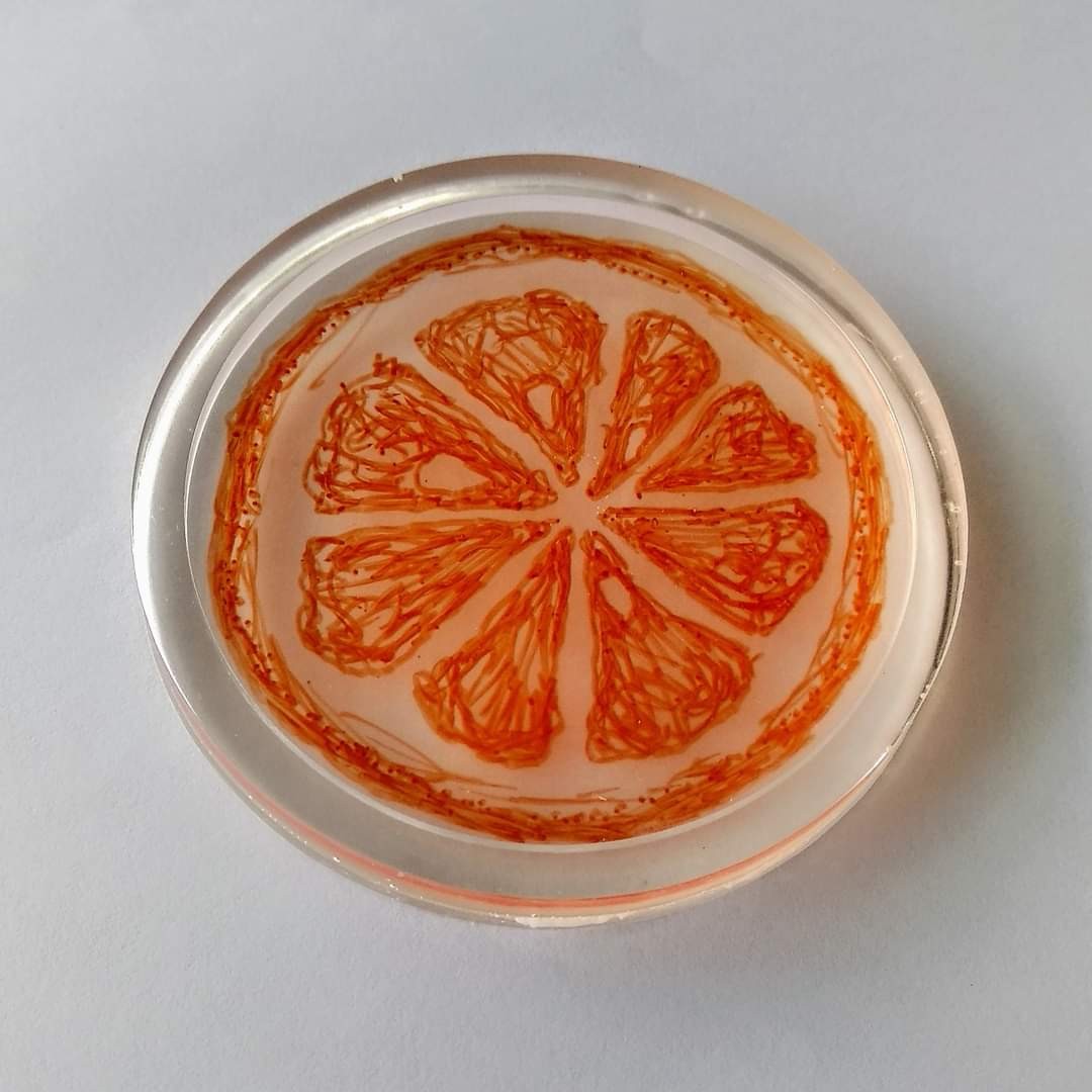Round clear resin coaster with a hand-drawn orange citrus cross-section — radiating segments in orange ink on pale pink