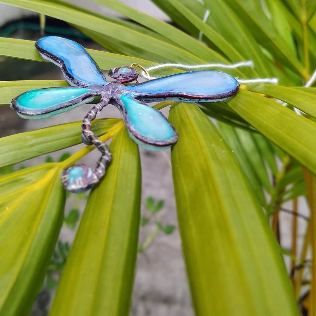 Blue resin dragonfly pendant with copper wire frame, photographed resting on tropical plant leaves
