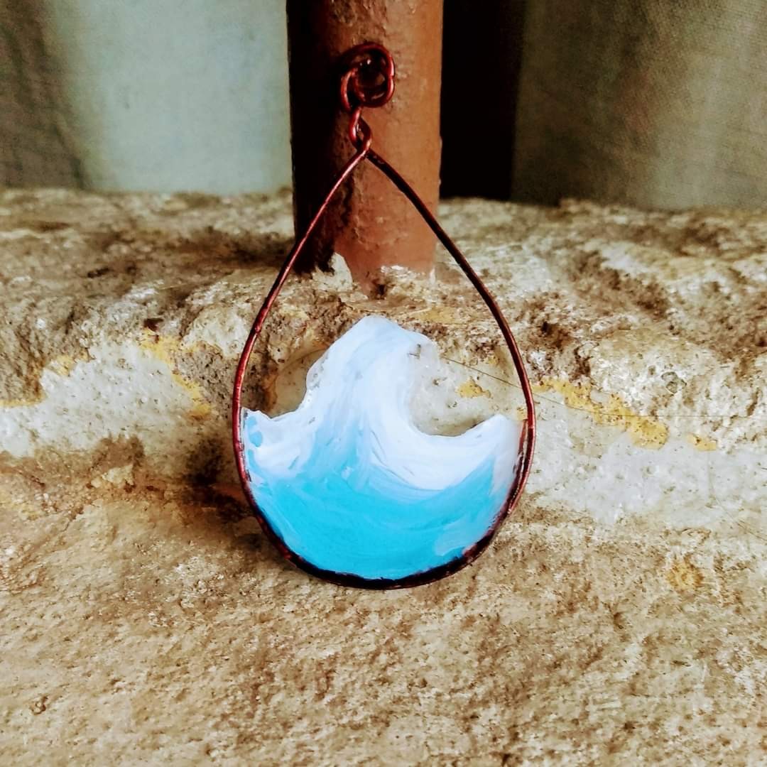 Teardrop copper wire pendant with a white and sky-blue resin wave inside, mounted against a rustic iron pipe