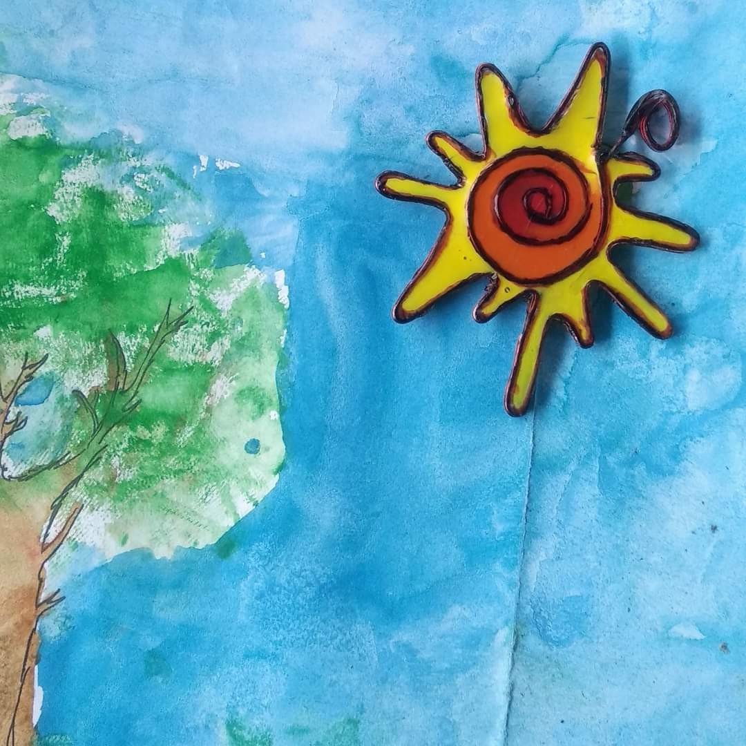 Wire-and-resin sun piece — yellow star shape with red spiral centre, displayed on a watercolour painting of a tree and blue sky