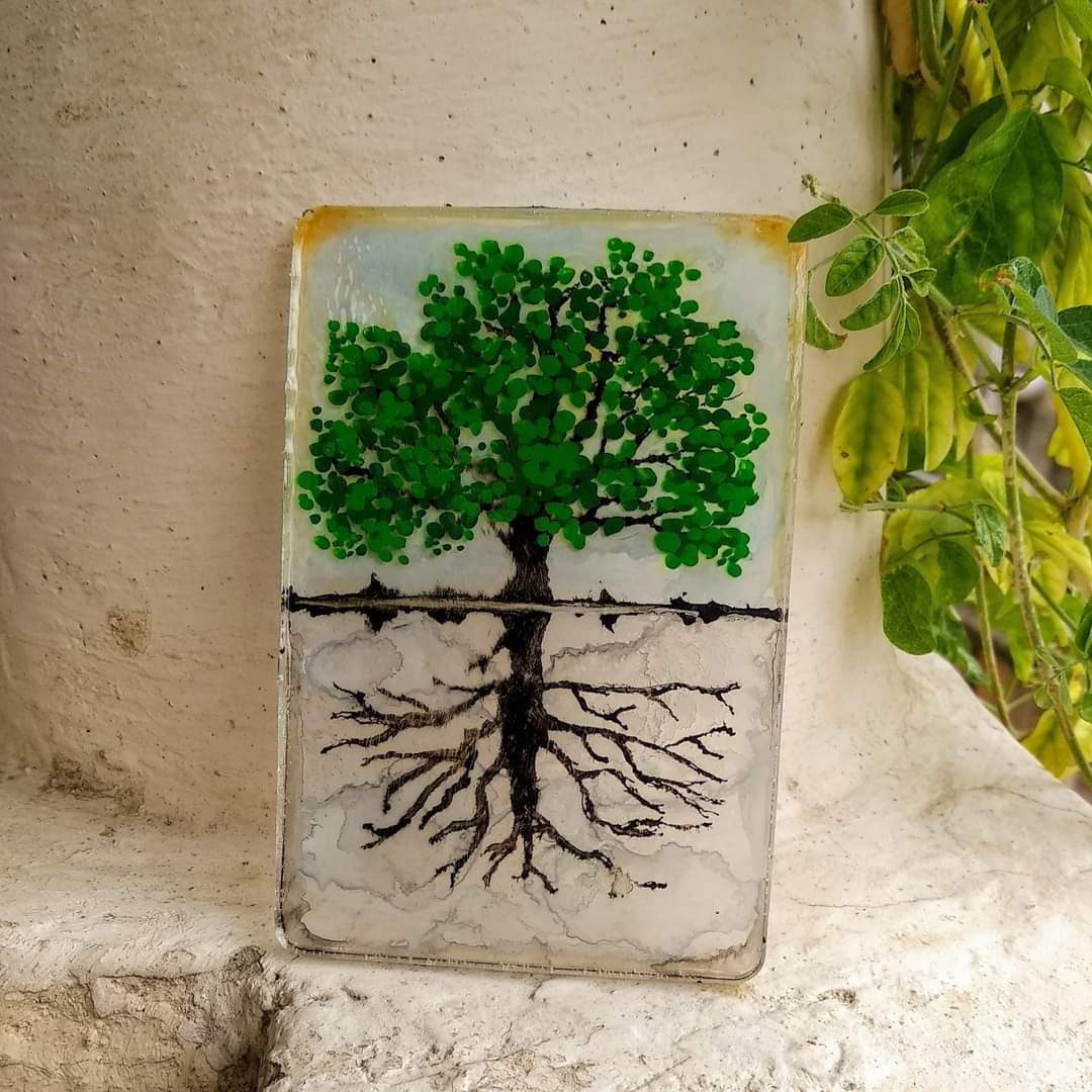 Vertical rectangular resin slab displayed against a white wall outdoors — green dotted-leaf tree canopy above soil line, detailed root system branching below